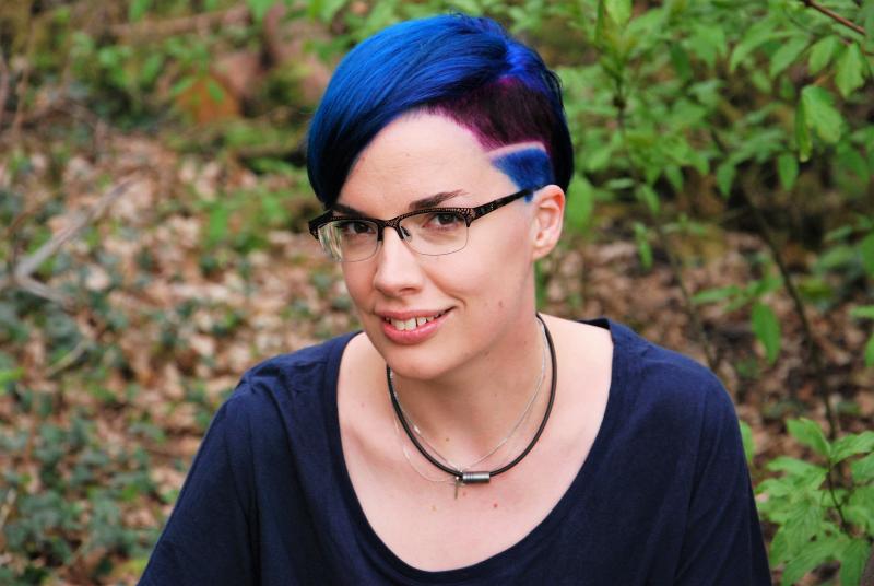 A picture of Marieke Nijkamp. She is looking at the camera and smiling. She has short, bright blue and pink hair, is wearing glasses, and is wearing a necklace.